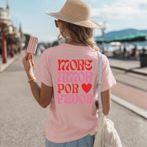 Peut inclure: T-shirt rose clair avec l'inscription "MORE AMOR POR FAVOR" en lettres rouges et roses, avec un cœur rouge. La personne porte un chapeau de paille et tient une glace.