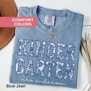 May include: Blue jean Comfort Colors t-shirt with "KINDER GARDEN" in large, white and blue floral letters. The text "mrs. anderson" is below the word "GARDEN". A gold necklace with a pendant is on the shirt.