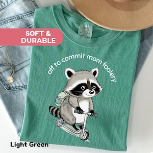 May include: Light green t-shirt featuring a raccoon on a scooter, with the words "off to commit mom foolery". The shirt also has the text "Soft & Durable" and "Comfort Colors" on the tag.