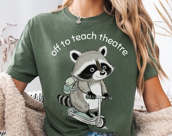 May include: A moss green t-shirt featuring a graphic of a cartoon raccoon wearing a backpack and riding a scooter. Above the raccoon, the white text reads "off to teach theatre" in a curved font. The shirt has short sleeves and a crew neckline.