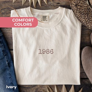 May include: An ivory short-sleeve t-shirt with a crew neckline featuring the embroidered brown text 1986 centered on the chest. The inner neck label displays the text Comfort Colors with size L. The shirt is made of a solid, light-colored fabric.