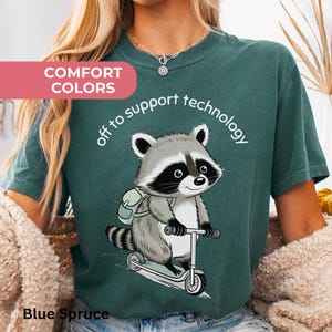 May include: Blue Spruce t-shirt with a cartoon raccoon riding a scooter, wearing a backpack, and the text "off to support technology." The shirt also has the text "Comfort Colors" and "Blue Spruce."
