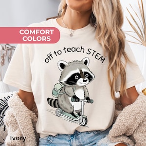 May include: Ivory-colored t-shirt with the text "off to teach STEM" and a cartoon raccoon riding a scooter. The raccoon has a backpack and is colored in shades of gray and black. The shirt also has the words "Comfort Colors" printed on it.