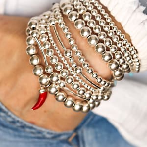 Può includere: Una collezione di braccialetti con perline d'argento di varie dimensioni, impilati su un polso. Un braccialetto presenta un piccolo ciondolo a forma di peperoncino rosso. I braccialetti sono indossati con un top bianco e denim blu.