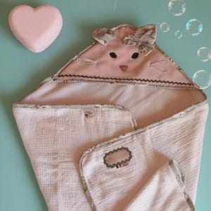 May include: A light pink hooded baby towel with a bunny face design, featuring black eyes and a bow. The towel is made of soft, textured fabric and has a decorative border. A pink heart-shaped bath bomb is also visible.
