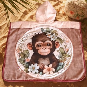 May include: A hooded baby towel featuring a charming monkey illustration surrounded by flowers. The towel is primarily pink and brown, with a circular design in the center. The towel has a hood and a dark pink border.