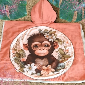 May include: Coral-colored hooded baby towel with a circular graphic of a brown monkey wearing a floral crown. The towel has a white border and is made of a soft, textured fabric. The monkey graphic is surrounded by flowers and leaves.