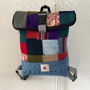 Peut inclure: Un sac à dos patchwork avec une fermeture à enroulement, composé de carrés de tissu en denim, rouge, vert, violet et motifs floraux. Le sac a des sangles grises et une boucle noire. Un petit logo rouge et blanc est visible à l'avant.