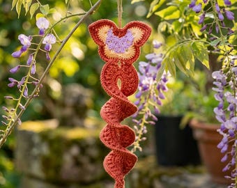 Può includere: Una decorazione all'uncinetto sospesa caratterizzata da una parte superiore a forma di farfalla color ruggine con un centro viola chiaro e una lunga coda a spirale color ruggine. L'oggetto è appeso a un filo sottile e presenta una trama in filato fatto a mano.