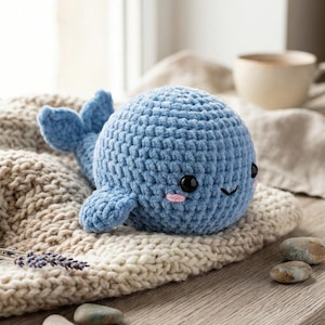 May include: A round, light blue crochet whale plush toy with a textured yarn surface. It features two shiny black plastic eyes, a small stitched black mouth, and two small pink embroidered patches on its cheeks to represent a blush effect.