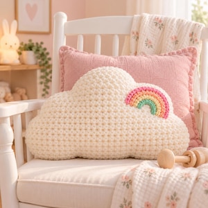 May include: A cream-colored, cloud-shaped crochet pillow featuring a small, multi-colored rainbow appliqué with pink, orange, yellow, and mint green yarn. The pillow is textured with a bobble stitch pattern and sits on a white wooden chair.