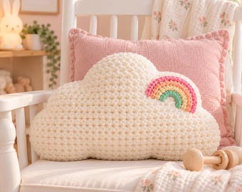Può includere: Un cuscino all'uncinetto a forma di nuvola color crema con una piccola applicazione arcobaleno realizzata con filati rosa, arancione, giallo e verde menta. Il cuscino presenta una trama a punto nocciolina e poggia su una sedia in legno bianco.