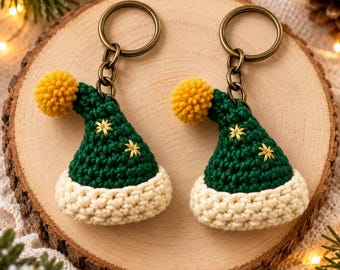 May include: Two crochet keychains shaped like miniature hats in dark green with a cream-colored brim, a yellow pom-pom at the tip, and small gold star embroidery. Each hat is attached to a bronze-toned metal split ring by a short chain link.