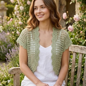 May include: A sage green crochet shrug featuring an openwork mesh pattern and a scalloped edge along the front opening and sleeves. The garment is worn over a simple white sleeveless dress, providing a lightweight layer with a textured finish.