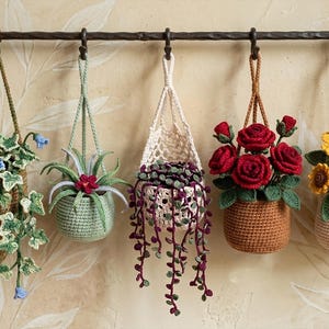 May include: Five crocheted hanging planters suspended from a dark metal rod. The set includes a cone-shaped ivy plant, a small green succulent, a white basket with trailing purple vines, a brown pot with red roses, and a beige pot with sunflowers.