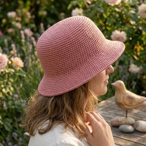 May include: A dusty rose bucket hat crafted with a dense, uniform crochet stitch pattern. The hat features a rounded crown and a short, slightly flared brim that encircles the head, providing a consistent texture throughout the entire accessory.