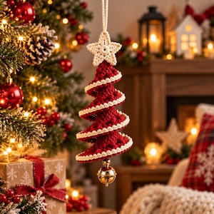 May include: A hanging Christmas tree ornament made of dark red crocheted yarn with cream-colored trim on the edges. The ornament features a crocheted star at the top and a small, shiny gold-colored metal bell hanging from the bottom point.