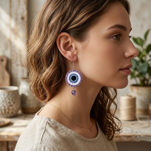 May include: A circular crochet earring featuring a concentric evil eye design in shades of black, blue, white, and purple. The earring hangs from a silver-toned hook and includes a small, dangling purple faceted bead at the bottom center.
