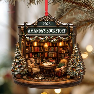 May include: A decorative hanging ornament shaped like a bookstore interior with the text AMANDA'S BOOKSTORE and 2026. It features two armchairs, a round table with an open book, a sleeping cat, bookshelves, and two Christmas trees with lights.