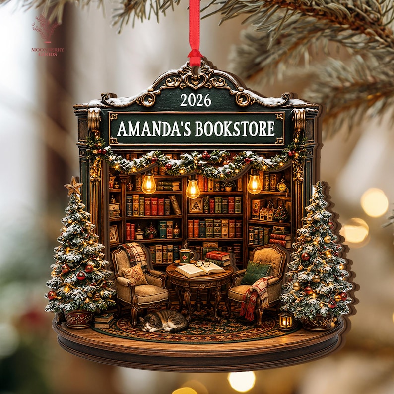 May include: A decorative hanging ornament shaped like a bookstore interior with the text AMANDA'S BOOKSTORE and 2026. It features two armchairs, a round table with an open book, a sleeping cat, bookshelves, and two Christmas trees with lights.