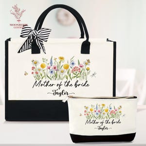 May include: A white tote bag and matching cosmetic bag with a black base and handles. Both bags feature a floral design with butterflies and dragonflies. The text "Mother of the bride Taylor" is printed on both bags. A black and white striped bow adorns the tote bag.