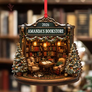 May include: A decorative hanging ornament shaped like a bookstore interior with the text AMANDA'S BOOKSTORE and 2026. It features two armchairs, a table with an open book, a sleeping cat, and two snow-dusted Christmas trees with warm lights.
