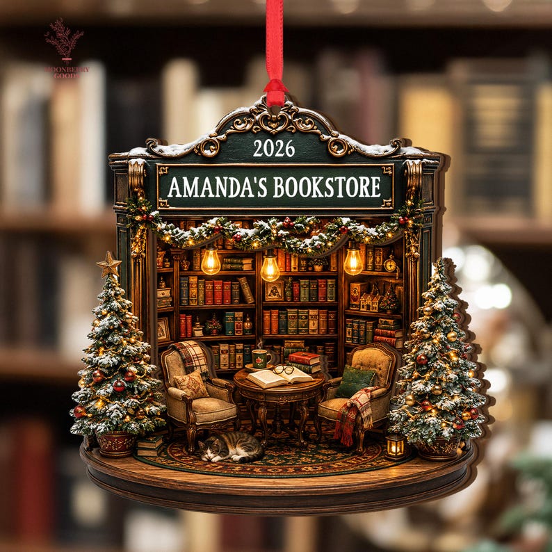 May include: A decorative hanging ornament shaped like a bookstore interior with the text AMANDA'S BOOKSTORE and 2026. It features two armchairs, a table with an open book, a sleeping cat, and two snow-dusted Christmas trees with warm lights.