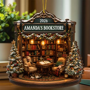 May include: A decorative hanging ornament featuring a miniature bookstore scene with the text 2026 and AMANDA'S BOOKSTORE. The scene includes two armchairs, a table with an open book, a cat, and two snow-dusted Christmas trees with warm lights.