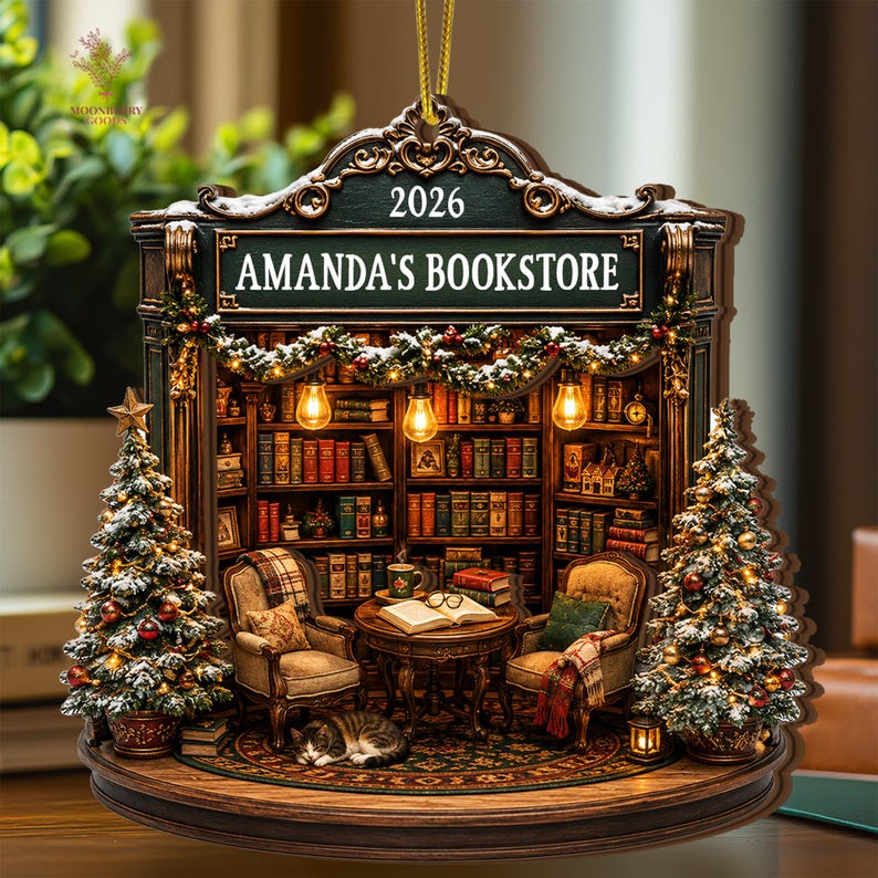 May include: A decorative hanging ornament featuring a miniature bookstore scene with the text 2026 and AMANDA'S BOOKSTORE. The scene includes two armchairs, a table with an open book, a cat, and two snow-dusted Christmas trees with warm lights.