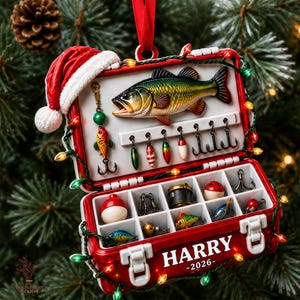 May include: A red tackle box Christmas ornament featuring a Santa hat, a bass fish relief, and various fishing lures. The front is personalized with the white text HARRY 2026. The box is wrapped in a string of small, glowing multicolored lights.