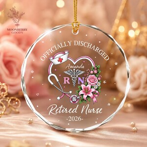 May include: A circular, faceted glass ornament featuring a gold hanging cord. The design includes a pink stethoscope, a nurse cap, a caduceus symbol, and pink flowers. Text reads: OFFICIALLY DISCHARGED, Amanda, RN, Retired Nurse, and -2026-.