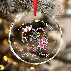 May include: A circular glass ornament with a red ribbon, featuring the text "OFFICIALLY DISCHARGED" and "Retired Nurse -2026-". The design includes a heart-shaped stethoscope, a nurse cap, a caduceus symbol, the name "Amanda", and pink flowers.