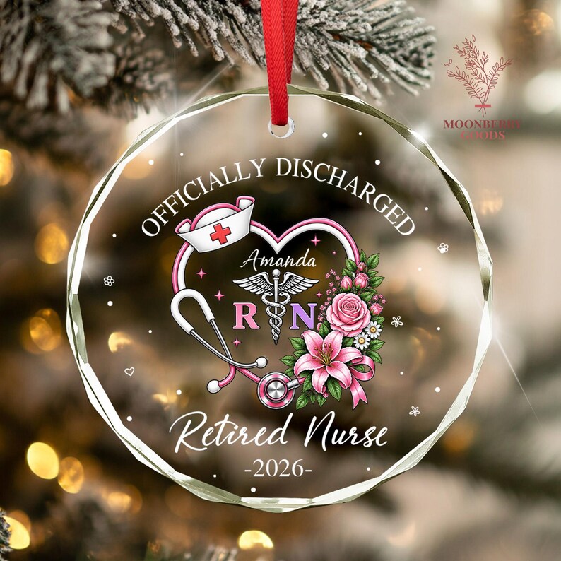 May include: A circular glass ornament with a red ribbon, featuring the text "OFFICIALLY DISCHARGED" and "Retired Nurse -2026-". The design includes a heart-shaped stethoscope, a nurse cap, a caduceus symbol, the name "Amanda", and pink flowers.