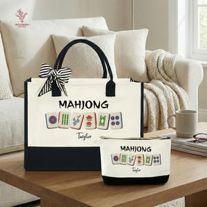 May include: A canvas tote bag and matching zippered pouch, both featuring the word "MAHJONG" and colorful tile illustrations. The tote has black handles and a black and white striped bow. The bottom of both items is black.