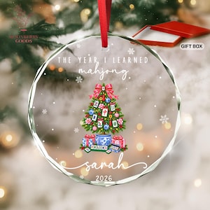 May include: A circular clear glass Christmas ornament with a red ribbon hanger. The design features a Christmas tree decorated with mahjong tiles, a red bow, and gifts at the base. Text reads: THE YEAR I LEARNED mahjong. sarah 2026.