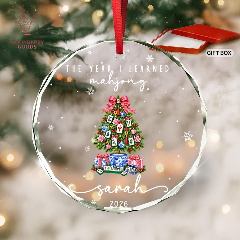 May include: A circular clear glass Christmas ornament with a red ribbon hanger. The design features a Christmas tree decorated with mahjong tiles, a red bow, and gifts at the base. Text reads: THE YEAR I LEARNED mahjong. sarah 2026.