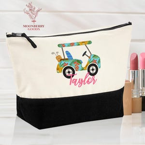 Op de afbeelding: Canvas make-up tas met een zwarte basis en een wit bovendeel. De tas heeft een kleurrijk golfkarretje ontwerp met de naam "Taylor" in roze script. De tas heeft een zwarte rits en een zwarte ritstrekker.