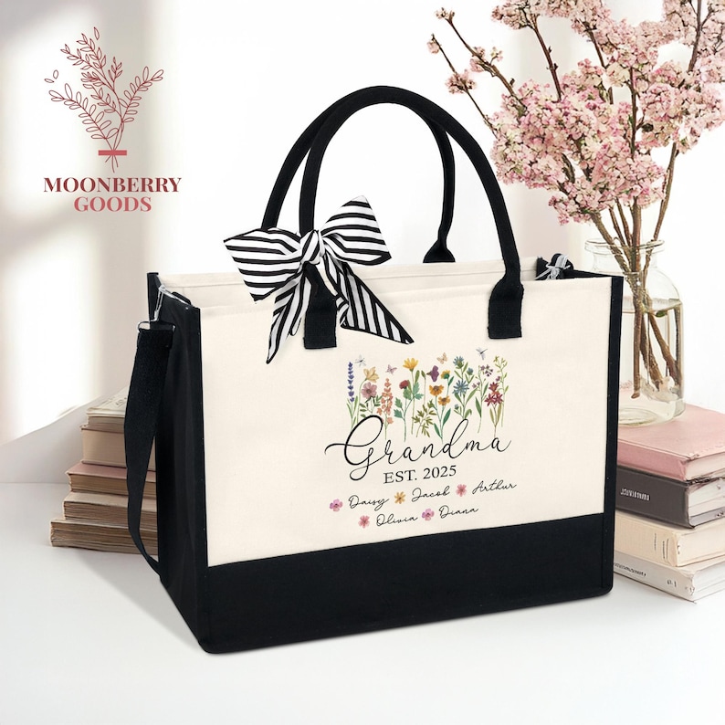 Personalized Grandma canvas tote bag with wildflower design, grandkids names and EST year printed, black handles, striped bow accent, cream and black color block, styled with books and pink flowers.