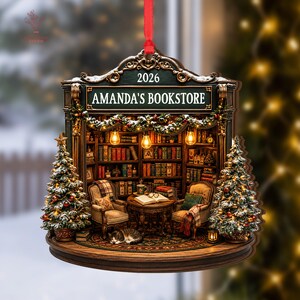 May include: A decorative hanging ornament shaped like a bookstore interior with a red ribbon. The sign reads AMANDA'S BOOKSTORE 2026. It features two armchairs, a table with an open book, a sleeping cat, and two Christmas trees with lights.