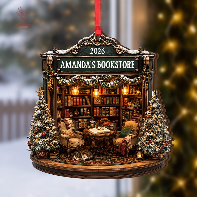 May include: A decorative hanging ornament shaped like a bookstore interior with a red ribbon. The sign reads AMANDA'S BOOKSTORE 2026. It features two armchairs, a table with an open book, a sleeping cat, and two Christmas trees with lights.