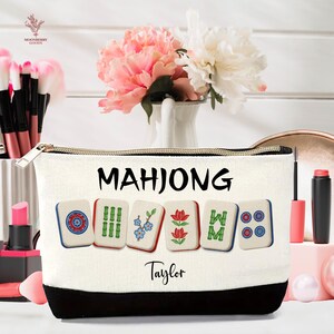 May include: A white canvas cosmetic bag with a black base and gold zipper. The bag features the word "MAHJONG" in black, along with images of Mahjong tiles. The name "Taylor" is printed below the tiles.