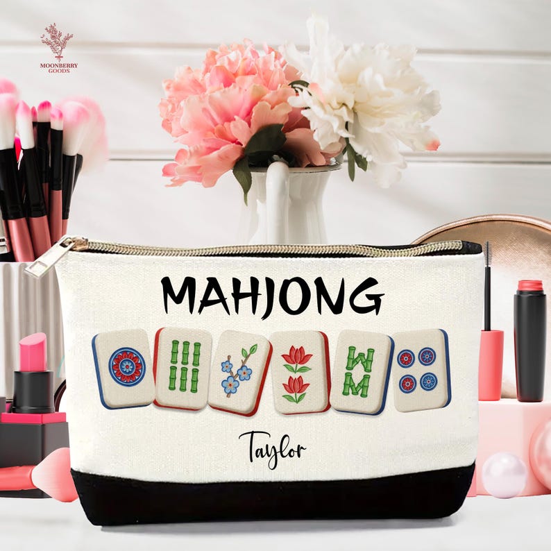 May include: A white canvas cosmetic bag with a black base and gold zipper. The bag features the word "MAHJONG" in black, along with images of Mahjong tiles. The name "Taylor" is printed below the tiles.