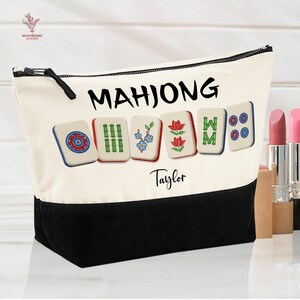 May include: Cream-colored canvas cosmetic bag with a black base and zipper. The bag features the word "MAHJONG" in black, along with colorful Mahjong tiles. The name "Taylor" is printed below the tiles. The bag is designed for storing makeup and other beauty products.