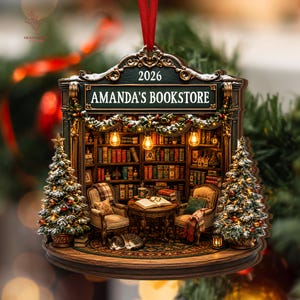 May include: A decorative Christmas ornament shaped like a bookstore interior with the text 2026 and AMANDA'S BOOKSTORE. It features two armchairs, a table with an open book, a sleeping cat, bookshelves, and two snow-dusted Christmas trees.