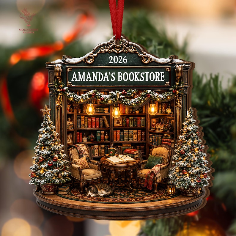 May include: A decorative Christmas ornament shaped like a bookstore interior with the text 2026 and AMANDA'S BOOKSTORE. It features two armchairs, a table with an open book, a sleeping cat, bookshelves, and two snow-dusted Christmas trees.