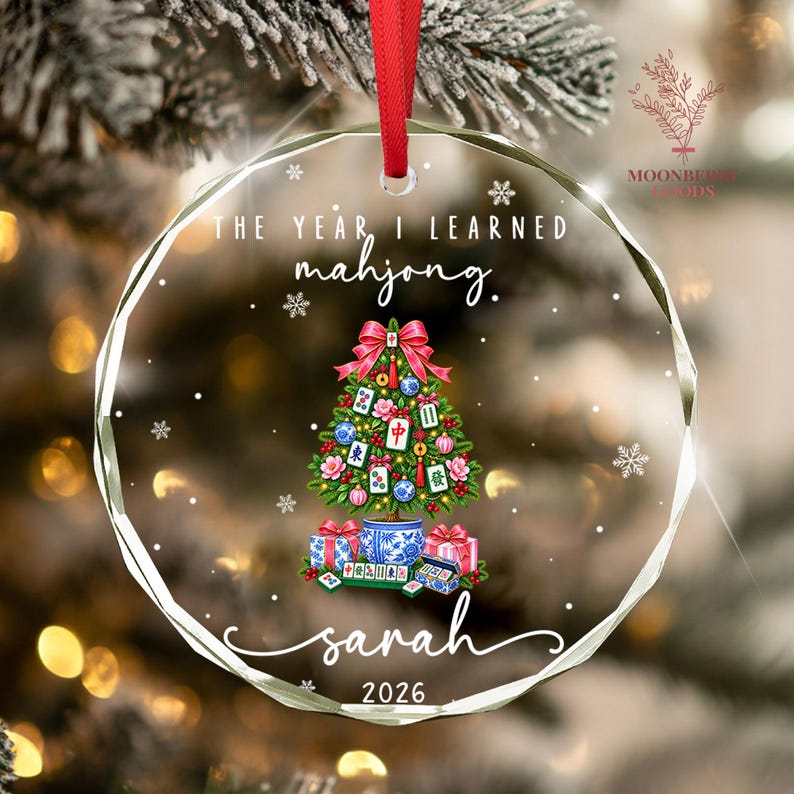 May include: A round, clear acrylic Christmas ornament with a red ribbon hanger. It features the text "THE YEAR I LEARNED mahjong." above a decorated tree with mahjong tiles, a blue and white planter base, gifts, the name "sarah", and "2026".