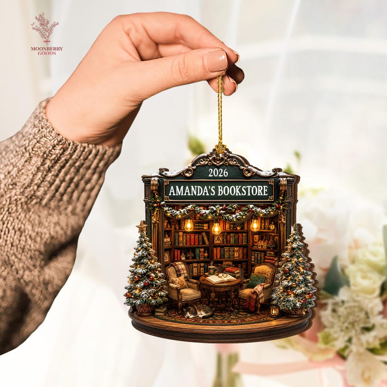 May include: A decorative hanging ornament shaped like a bookstore storefront with the text AMANDA'S BOOKSTORE and 2026. The design features a detailed interior with bookshelves, two armchairs, a small table, a cat, and two decorated Christmas trees.
