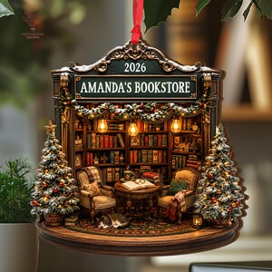 May include: A decorative hanging ornament shaped like a bookstore interior with the text AMANDA'S BOOKSTORE and 2026. It features two armchairs, a round table with an open book, a sleeping cat, bookshelves, and two Christmas trees with lights.