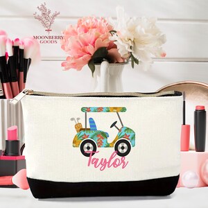 Op de afbeelding: Een witte make-uptas met een zwarte onderkant en een gouden rits. De tas heeft een golfkar-ontwerp met een tropisch bloemenpatroon en de naam "Taylor" in roze script. De tas is versierd met een golfkar-ontwerp.