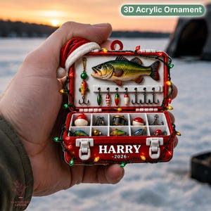 May include: A 3D acrylic ornament shaped like a red fishing tackle box with a Santa hat on top. The box contains a fish figure, various lures, and hooks. The front is printed with the name HARRY and the year 2026, decorated with string lights.